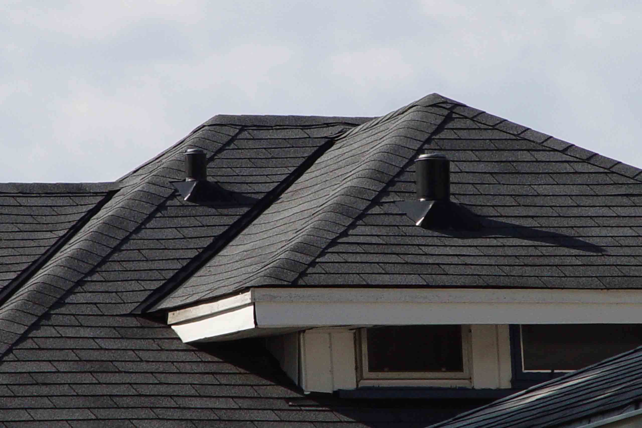 Close-up view of a house roof with dark asphalt shingles, two roof vents, and part of a dormer with white trim and windows under the eaves.