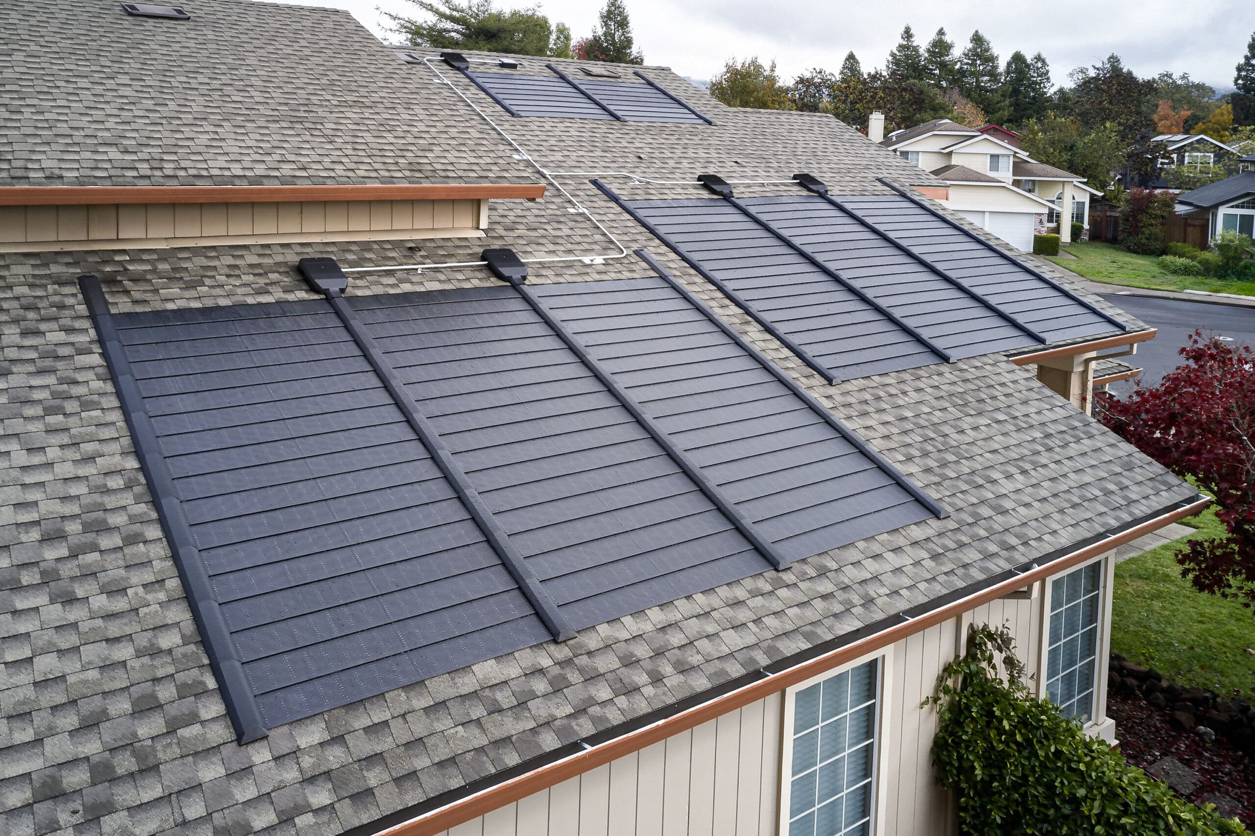 A house features a gray shingle roof with multiple black solar panels installed on two sections facing the street.