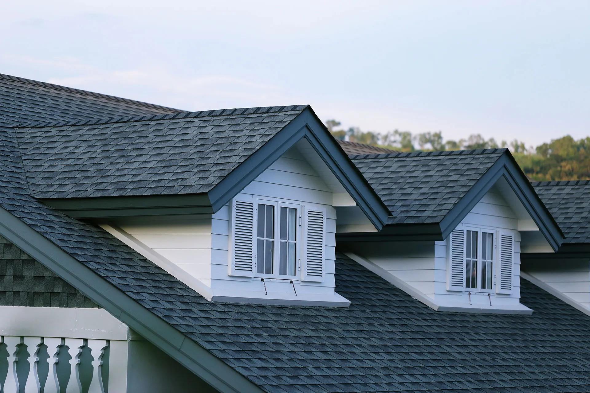 Two white dormer windows with shutters are built into a sloped roof covered with gray asphalt shingles. Trees are visible in the background.