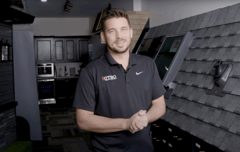 A man in a black polo shirt stands indoors next to a display featuring roof shingles and a skylight, smiling at the camera.