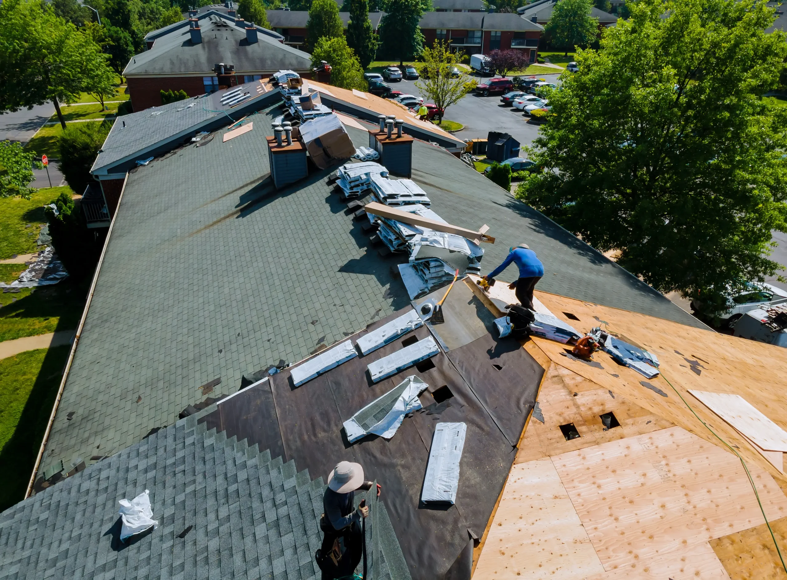 Aerial view of workers installing new shingles and roofing materials on a large residential building. Tools and supplies are spread across the roof.