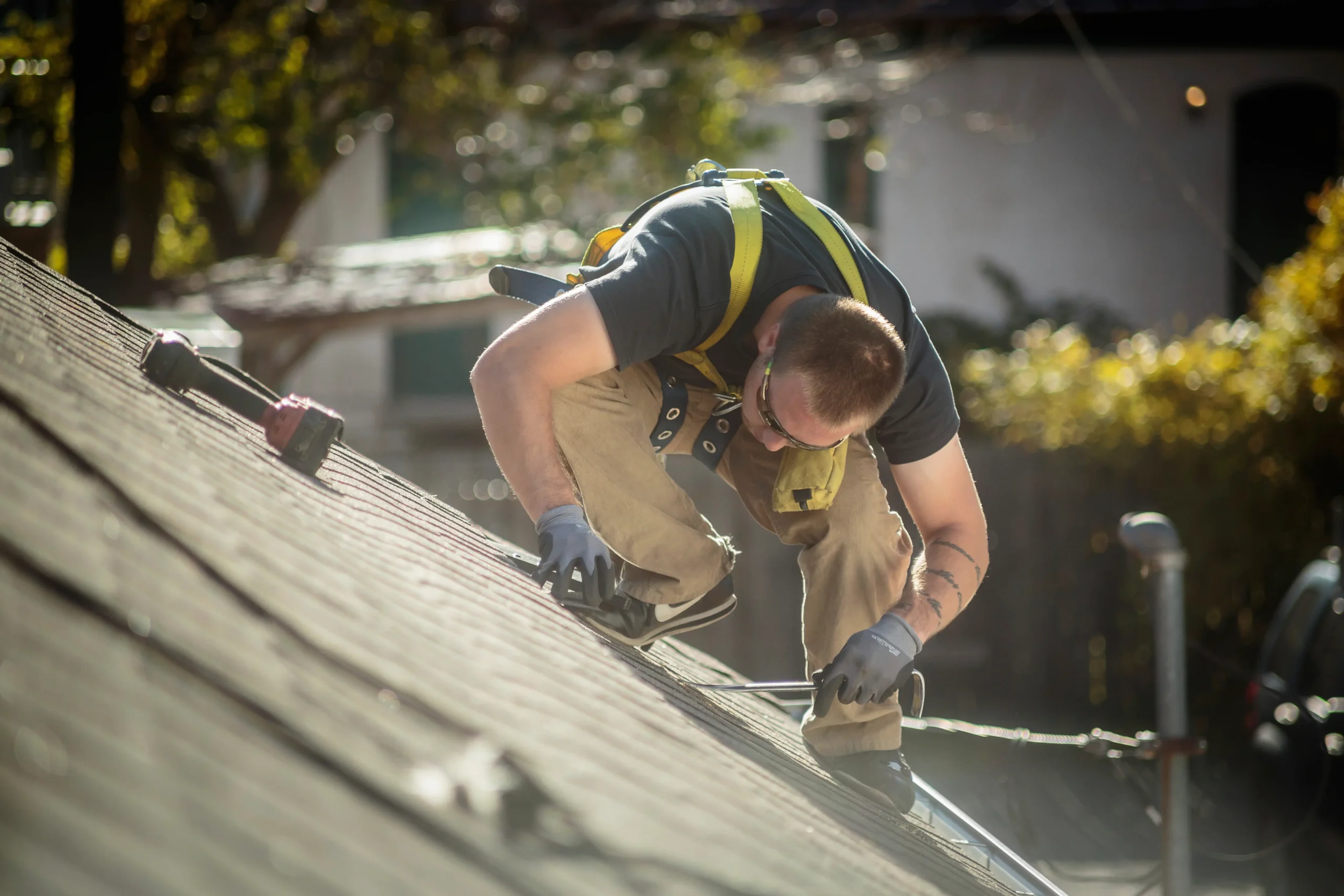 A person wearing safety gear kneels on a sloped roof while performing maintenance or repair work on a sunny day.