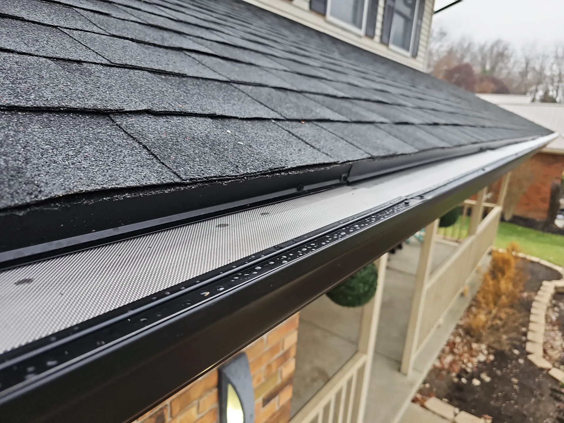 Close-up of a house's roof edge showing black shingles and a gutter covered with a fine mesh guard, with part of a porch and landscaping visible below.