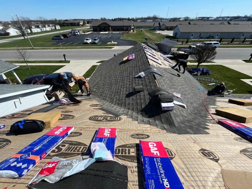 Roofers work on installing new shingles on a residential roof, with roofing materials and tools spread out across the surface and vehicles parked in the background.