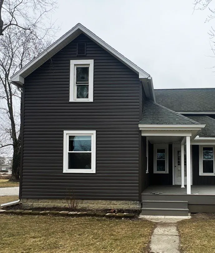 A dark-sided house with two white-framed windows vertically misaligned, grey porch, and bare yard, photographed on an overcast day.