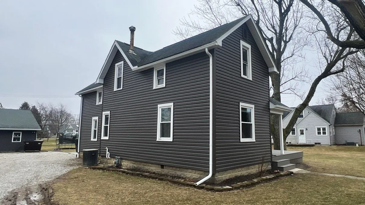 A two-story house with dark gray vinyl siding, multiple white-trimmed windows, and a small front porch sits on a grassy lot with a gravel driveway.
